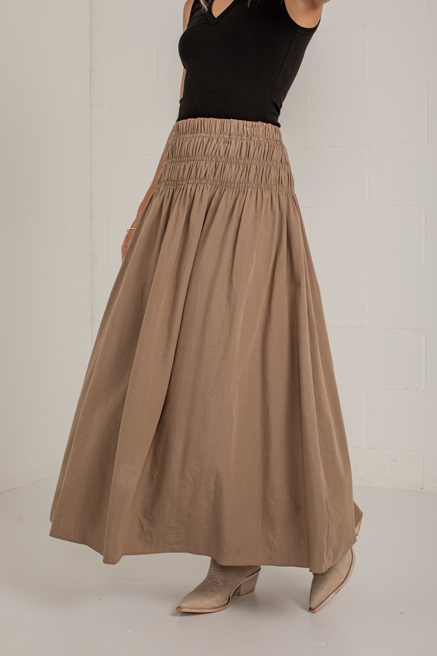 A woman wearing a high-waisted, pleated taupe-brown maxi skirt paired with a black sleeveless top and beige pointed-toe heeled mules, standing against a white brick wall.
