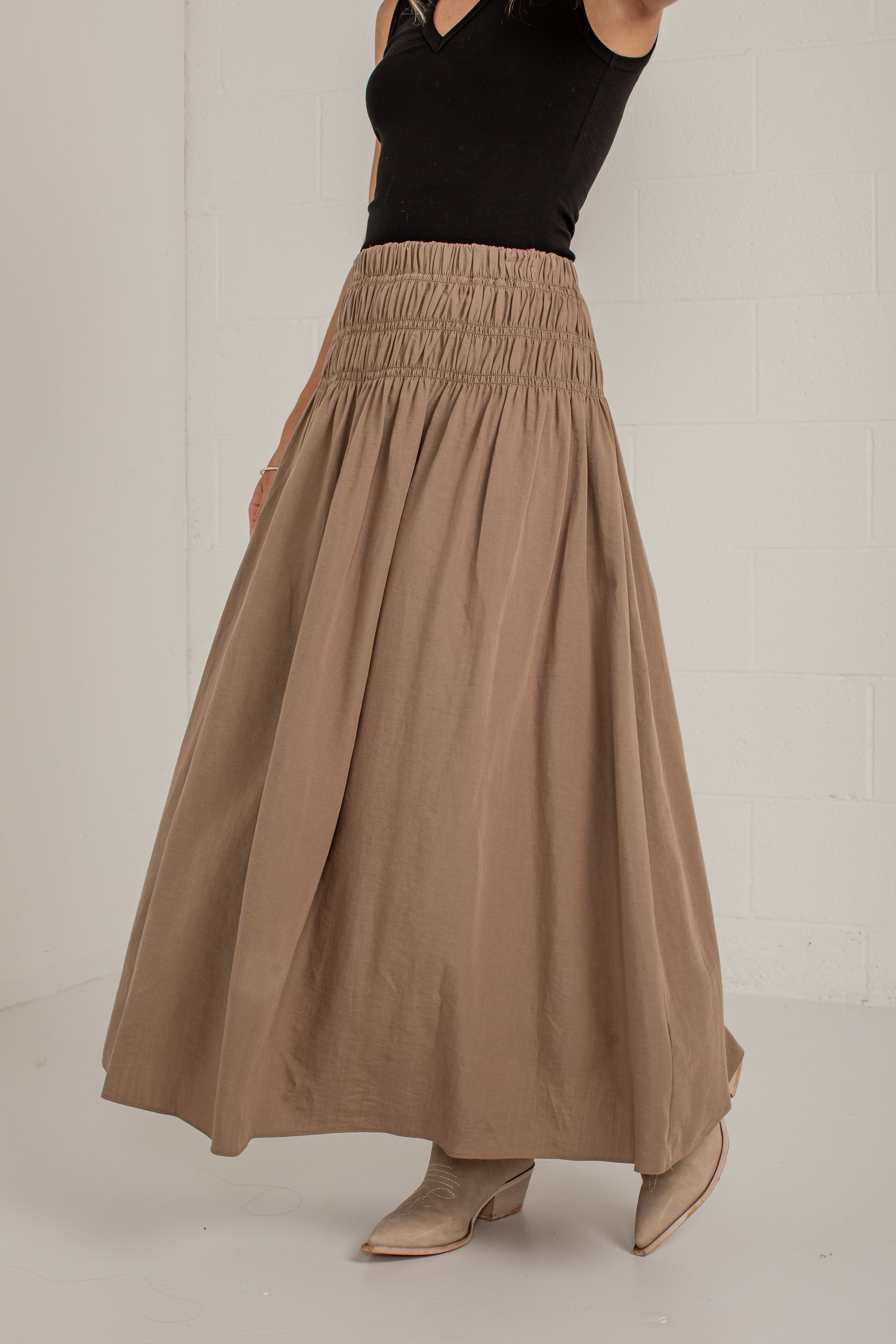 A woman wearing a high-waisted, pleated taupe-brown maxi skirt paired with a black sleeveless top and beige pointed-toe heeled mules, standing against a white brick wall.