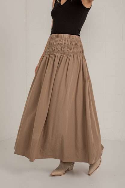 A woman wearing a high-waisted, pleated taupe-brown maxi skirt paired with a black sleeveless top and beige pointed-toe heeled mules, standing against a white brick wall.