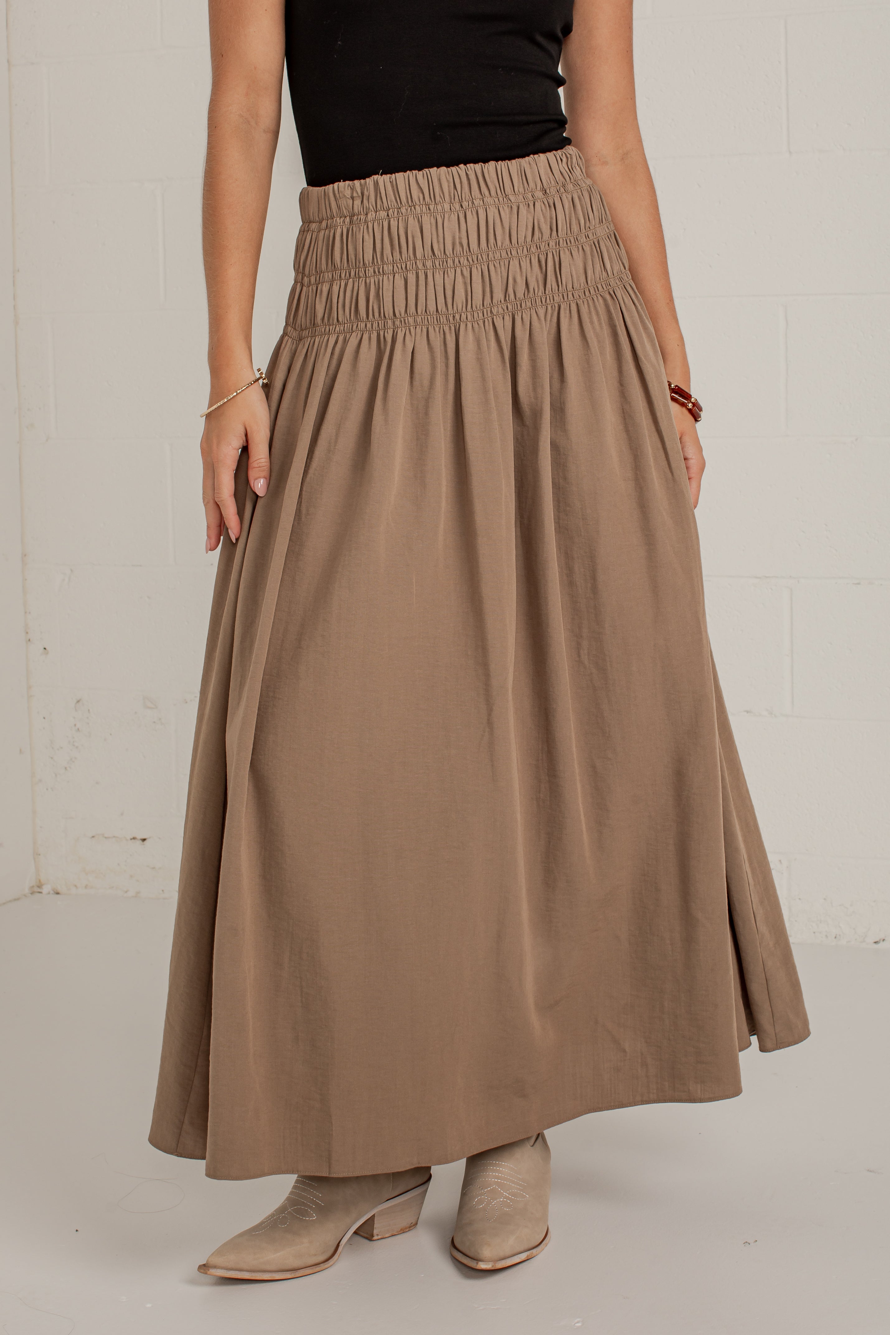 A woman wearing a high-waisted, smocked maxi skirt in a warm taupe-brown color, paired with a black top and beige heeled boots, standing against a white brick wall.
