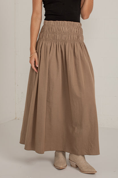 A model wearing a high-waisted, pleated maxi skirt in a warm taupe-brown color, paired with a black top and beige ankle boots, standing against a white brick wall.