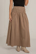 A model wearing a high-waisted, pleated maxi skirt in a warm taupe-brown color, paired with a black top and beige ankle boots, standing against a white brick wall.