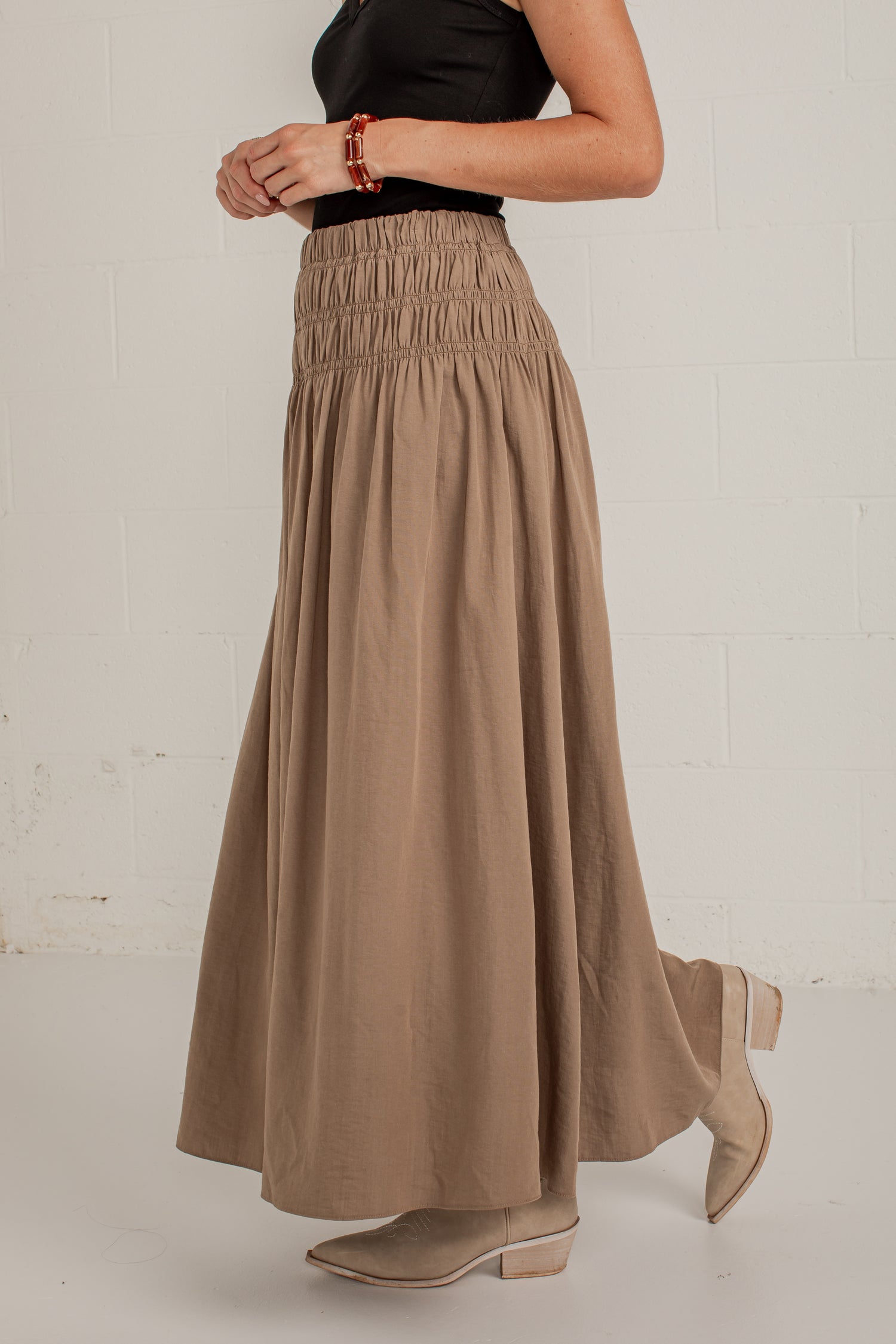 A woman wearing a high-waisted, long taupe-brown smocked maxi skirt with a black top and beige ankle boots, standing against a white brick wall.