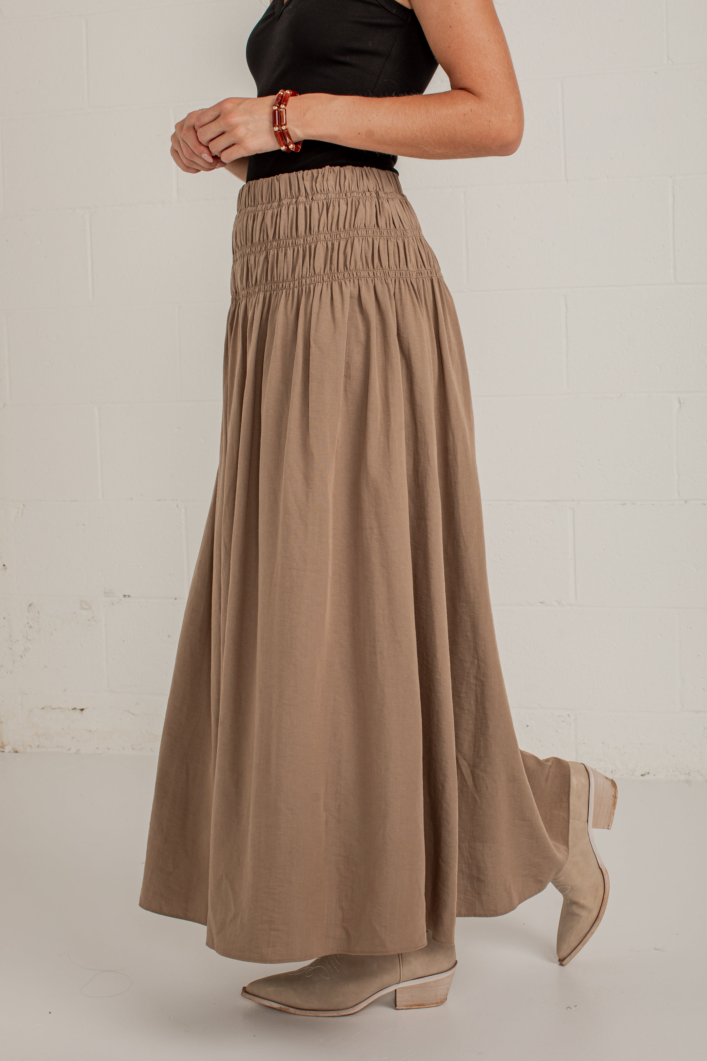 A woman wearing a high-waisted, long taupe-brown smocked maxi skirt with a black top and beige ankle boots, standing against a white brick wall.