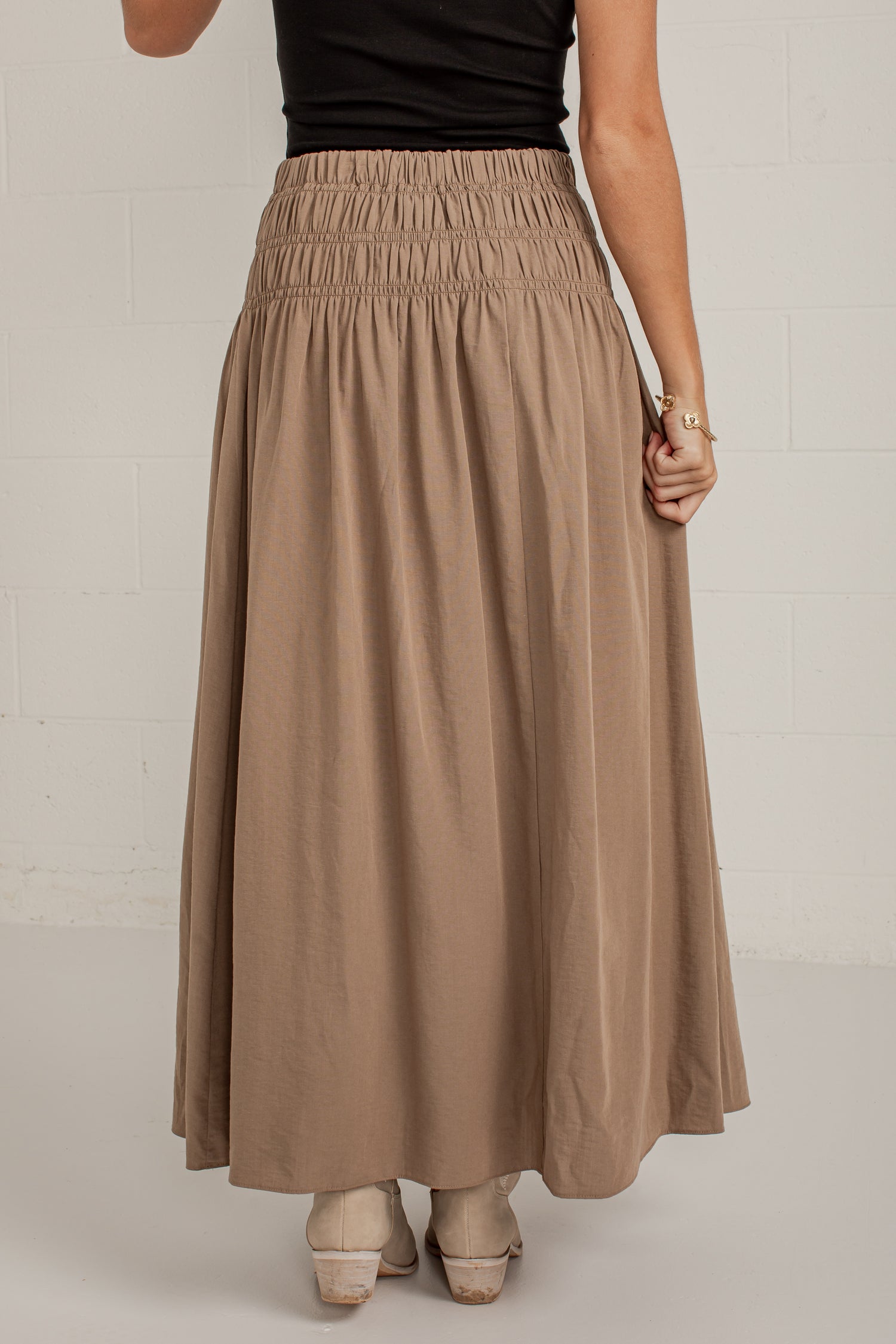 A woman wearing a long, flowing taupe-brown maxi skirt with an elastic waistband, paired with a black top and beige heels, standing against a white brick wall.