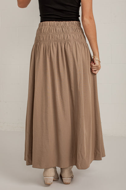 A woman wearing a long, flowing taupe-brown maxi skirt with an elastic waistband, paired with a black top and beige heels, standing against a white brick wall.
