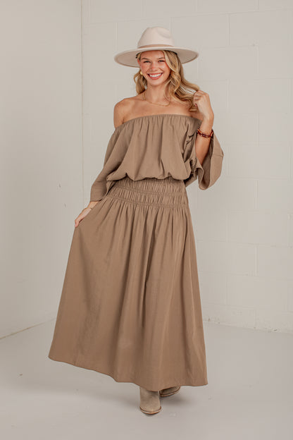 A woman modeling a taupe-brown off-the-shoulder maxi dress with a smocked waist, paired with a beige wide-brim hat and beige boots, standing against a white brick wall.