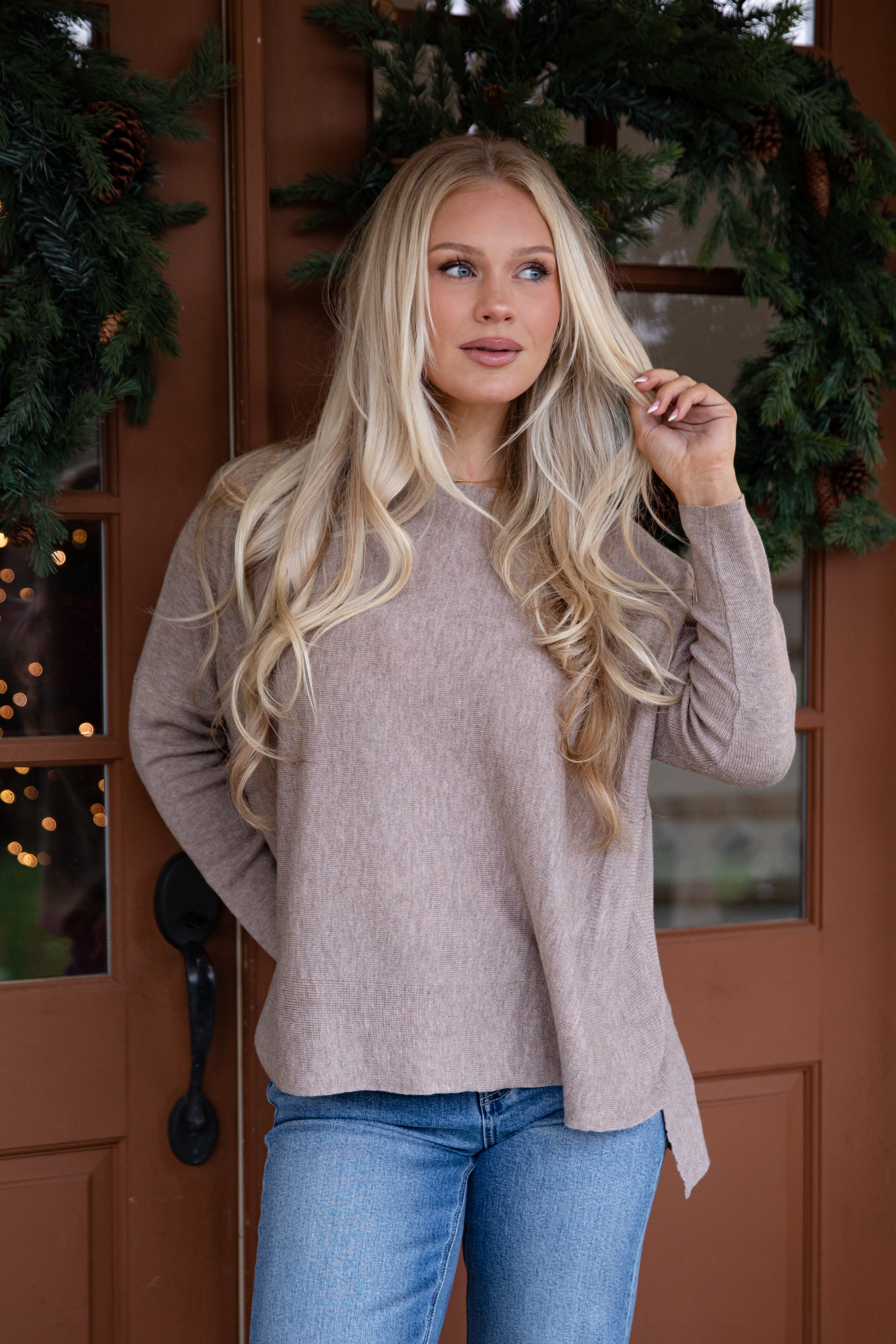 Woman wearing a beige sweater and blue jeans standing in front of a wooden door with decorative greenery.