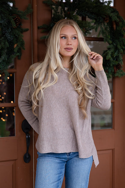 Woman wearing a beige sweater and blue jeans standing in front of a wooden door with decorative greenery.