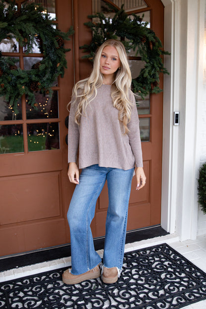 Woman standing in front of a brown door with Christmas decorations, wearing a beige sweater and blue jeans.