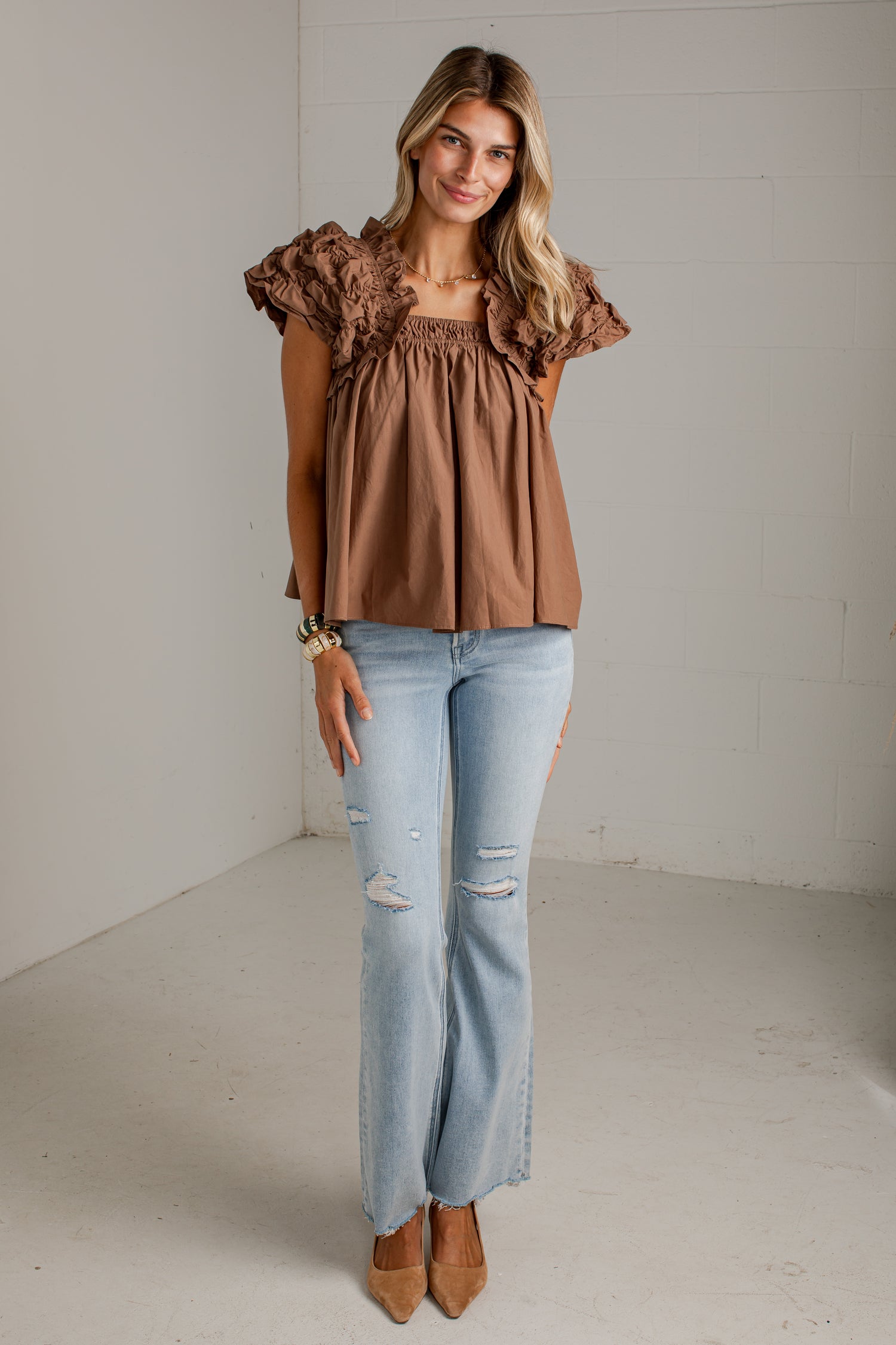 A woman modeling a brown ruffled short-sleeve top with a smocked bodice, paired with light-wash distressed jeans and tan pointed-toe heels, standing in a bright studio with white walls.
