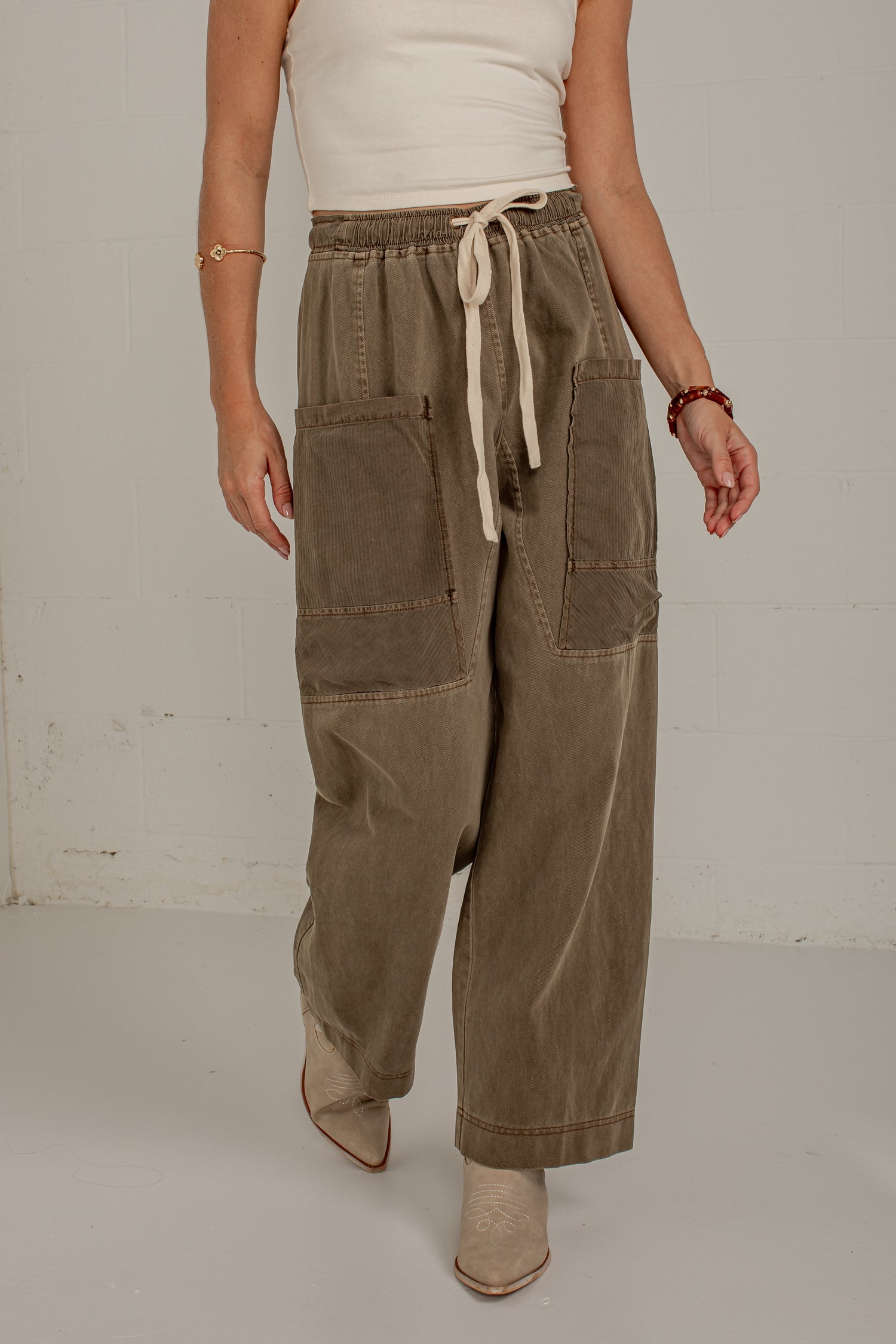 A model is shown from the waist down wearing wide-leg, cropped brown corduroy pants with a cream drawstring waist and large patch pockets. The background is a plain, light-colored studio wall.