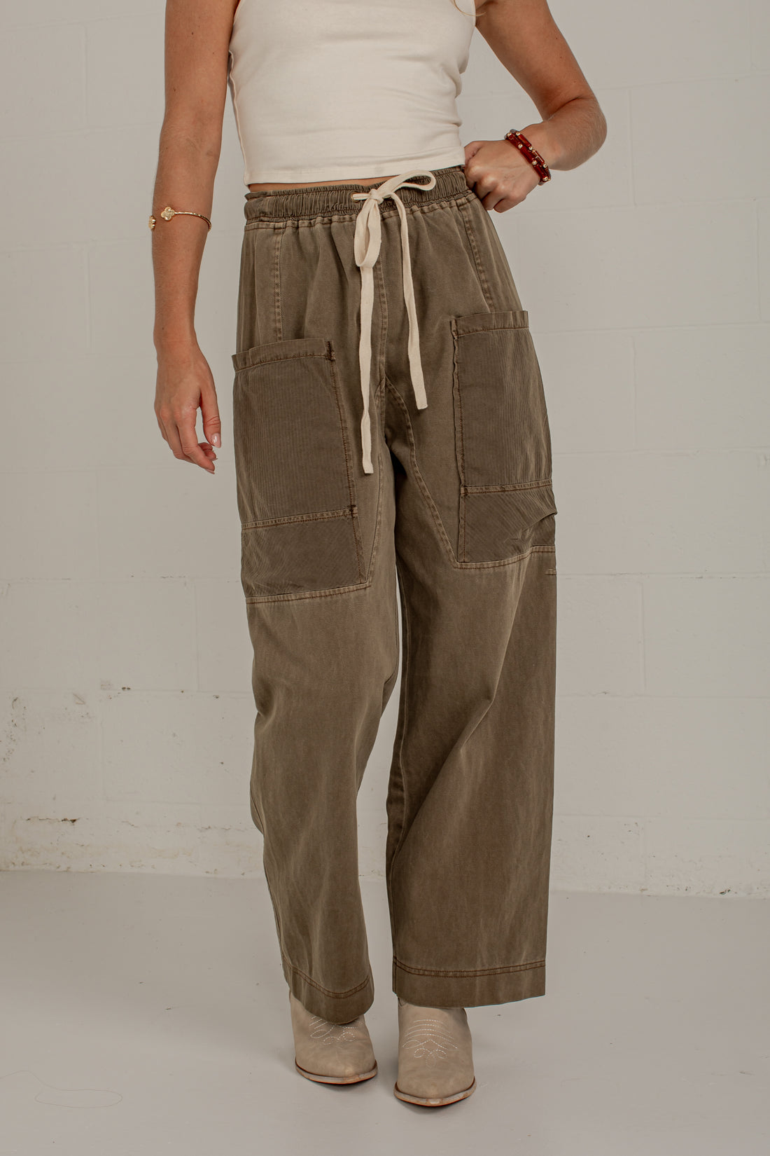 A person wearing wide-leg brown utility-style pants with a cream drawstring waist, large front patch pockets, and contrast paneling on the legs, paired with a white top and beige ankle boots against a plain light wall.