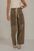 A person wearing wide-leg brown utility-style pants with a cream drawstring waist, large front patch pockets, and contrast paneling on the legs, paired with a white top and beige ankle boots against a plain light wall.