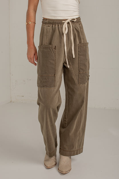 A person wearing wide-leg taupe corduroy pants with a cream drawstring waist, large patch pockets, and beige ankle boots, standing against a plain off-white studio wall.