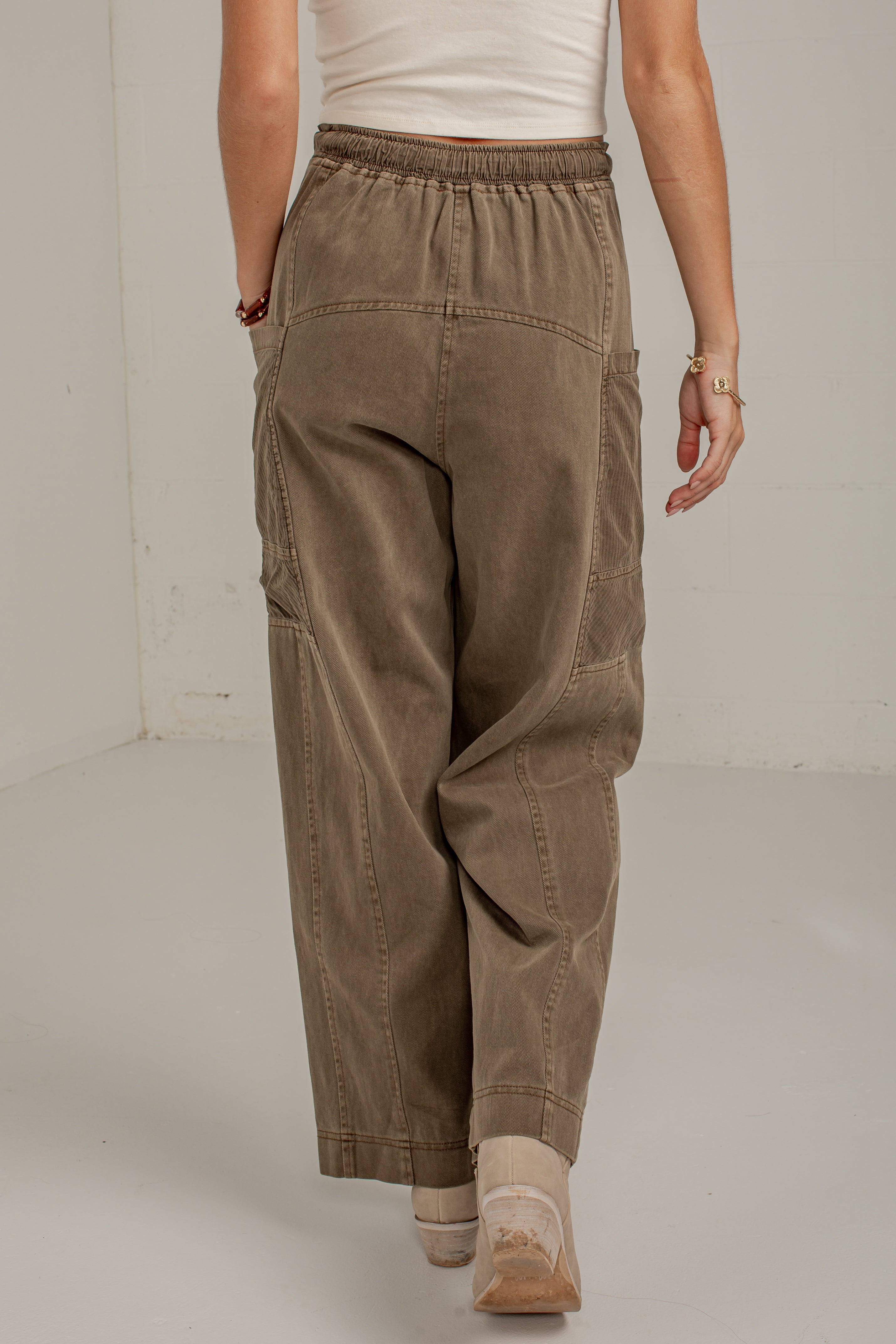 A person shown from the back wearing loose, wide-leg cargo pants in a muted taupe-brown color, paired with a white ribbed top and beige lace-up shoes, in a minimalist studio setting.