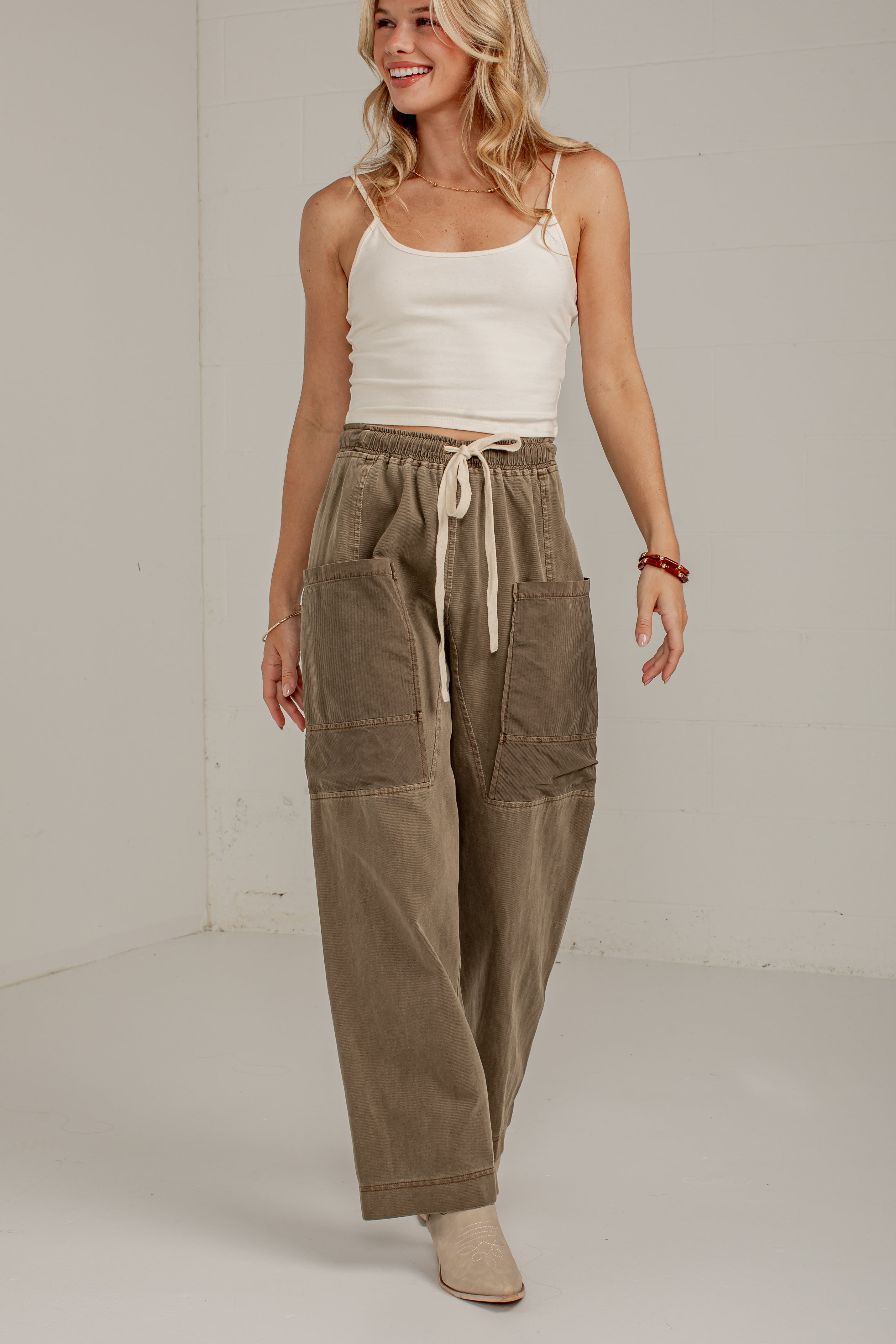 A woman modeling a pair of wide-leg, relaxed-fit cargo pants in a muted taupe-brown color. The pants feature an elastic drawstring waist and large front cargo pockets.