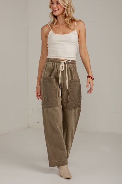 A woman modeling a pair of wide-leg, relaxed-fit cargo pants in a muted taupe-brown color. The pants feature an elastic drawstring waist and large front cargo pockets.