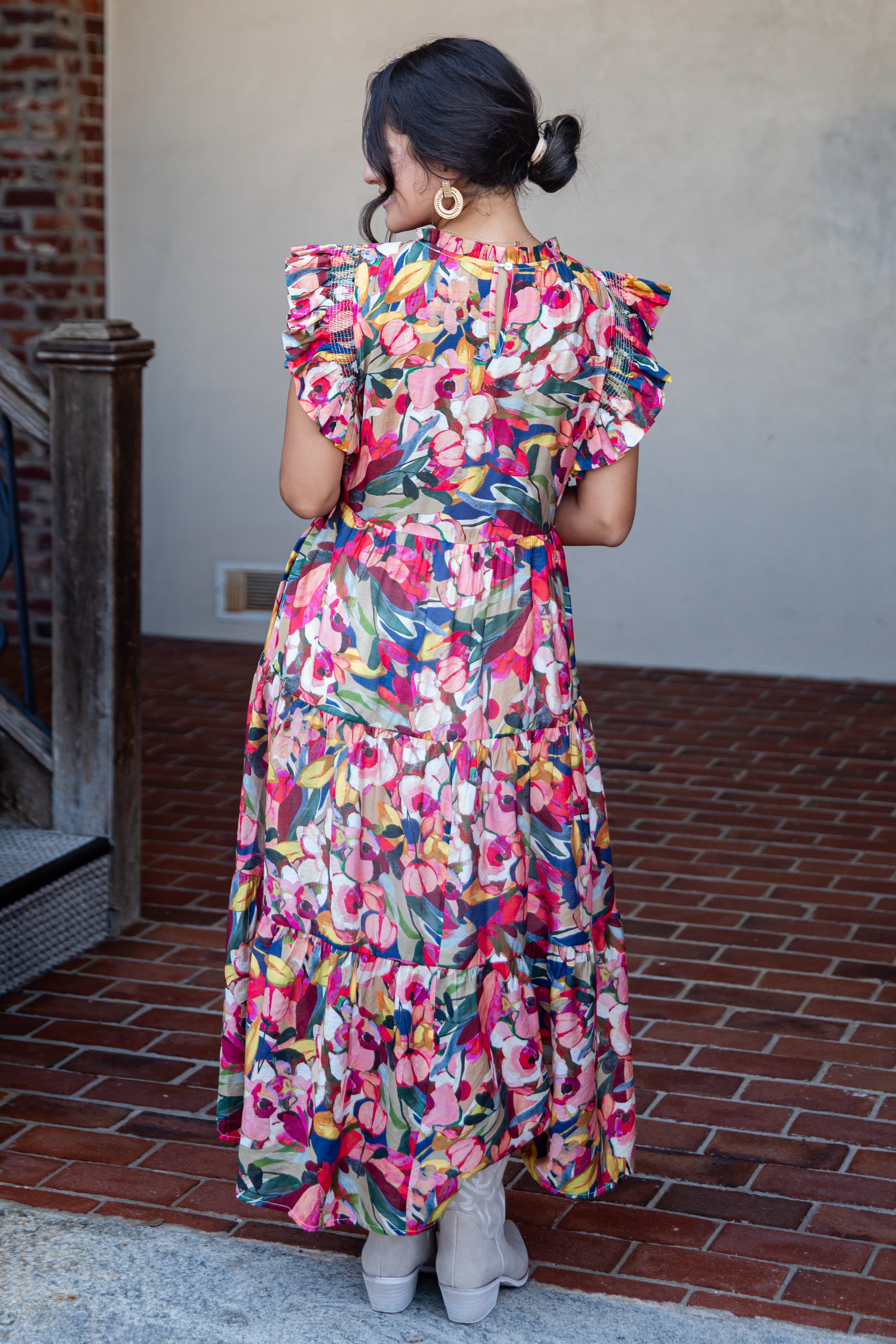 A woman viewed from behind wearing a colorful floral midi dress with ruffled short sleeves and a tiered skirt, standing on a brick patio against a stucco wall.