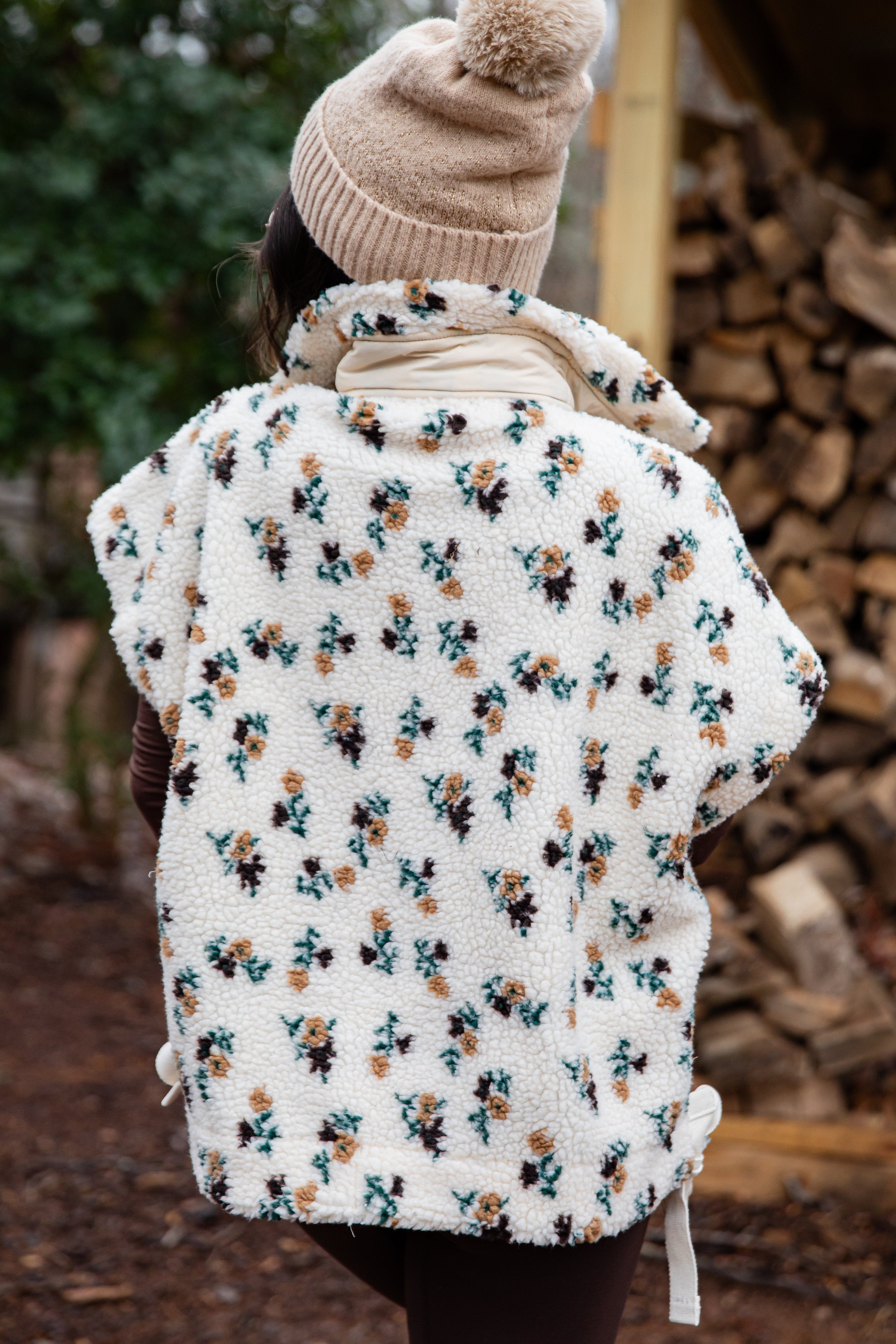 Person wearing a floral-patterned white fleece vest with a beanie in a forest setting.