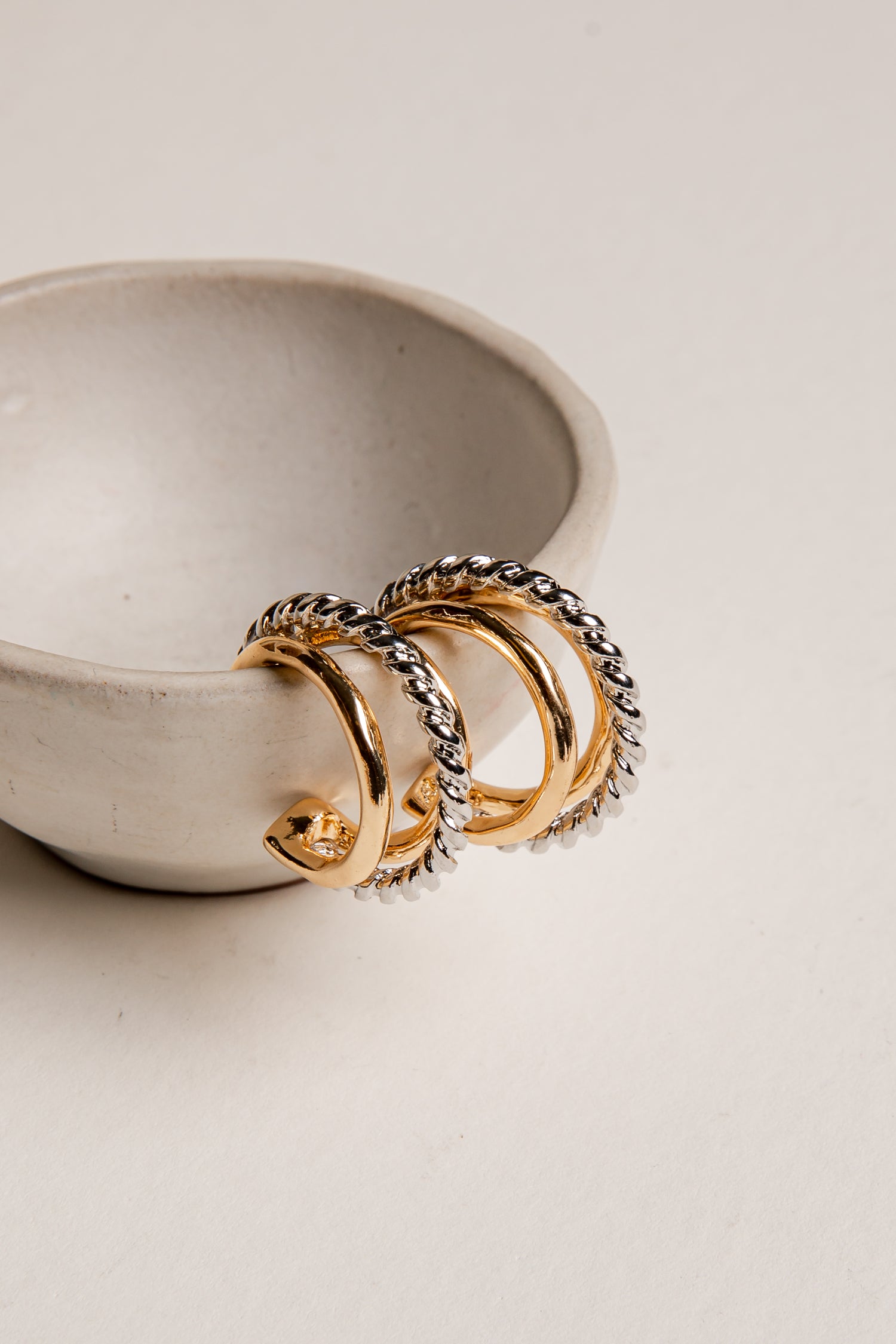 Gold and silver hoop earrings on a light beige surface with a ceramic bowl in the background.