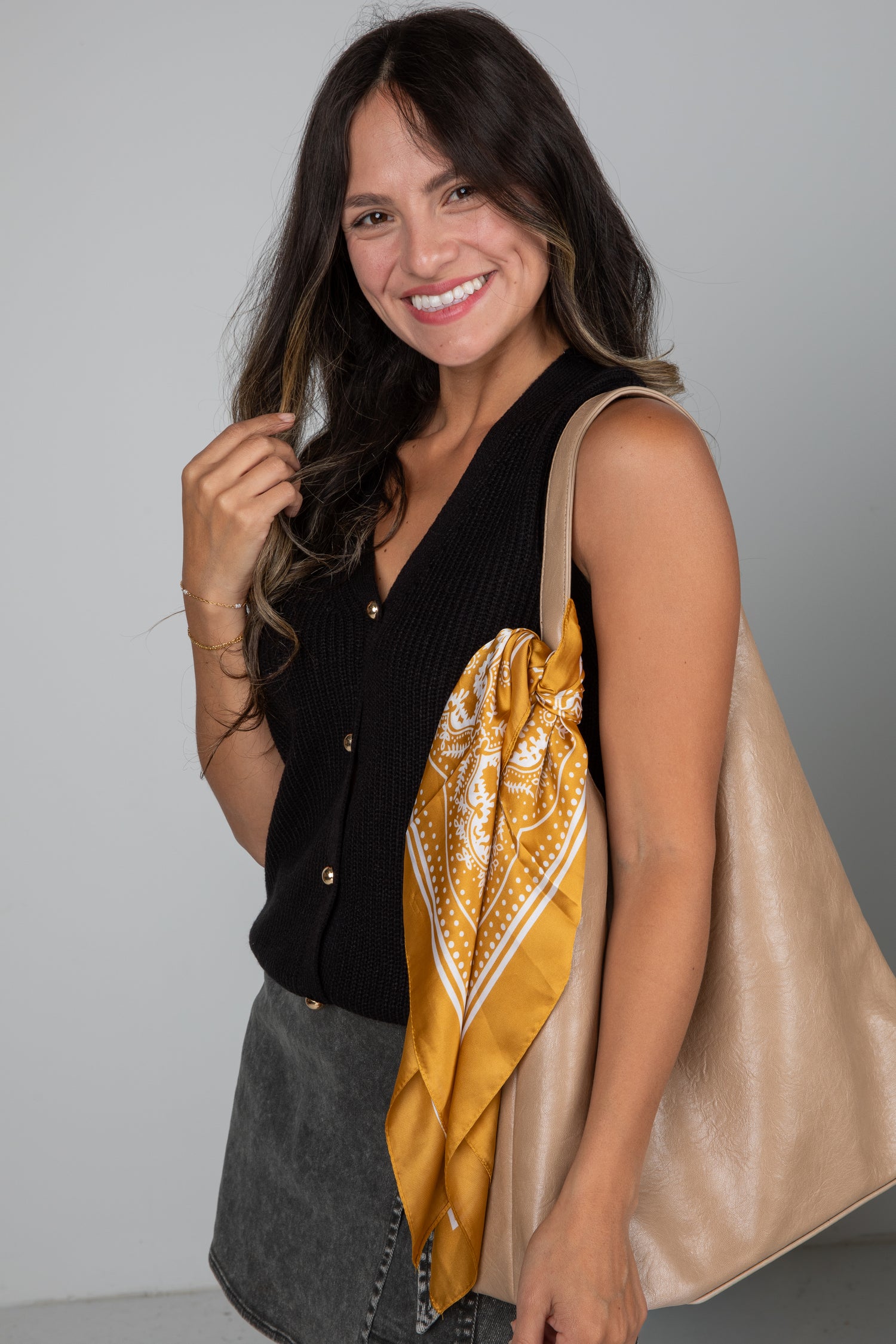 Woman holding a beige tote bag with a yellow patterned scarf over her shoulder against a gray background