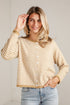 A woman wearing a beige and white striped button-up cardigan sweater with light blue jeans, posing against a white paneled wall.
