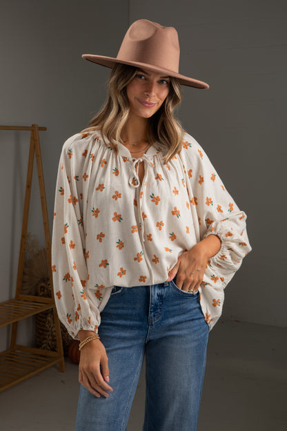 Woman wearing a floral blouse, blue jeans, and a brown hat in a neutral setting