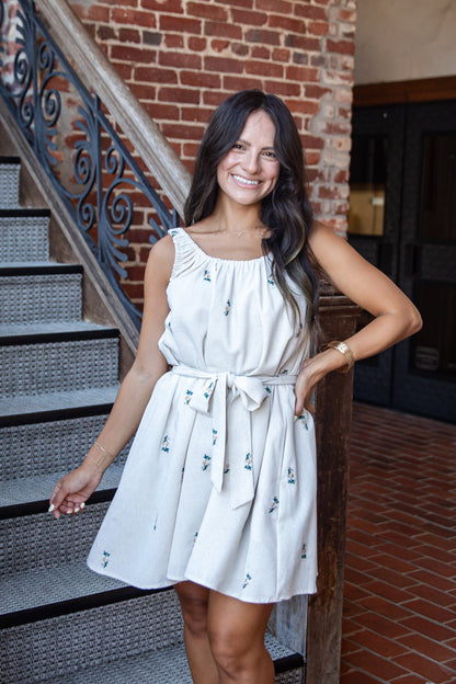 A woman wearing a sleeveless white mini dress with small green floral prints, standing in front of a brick staircase.