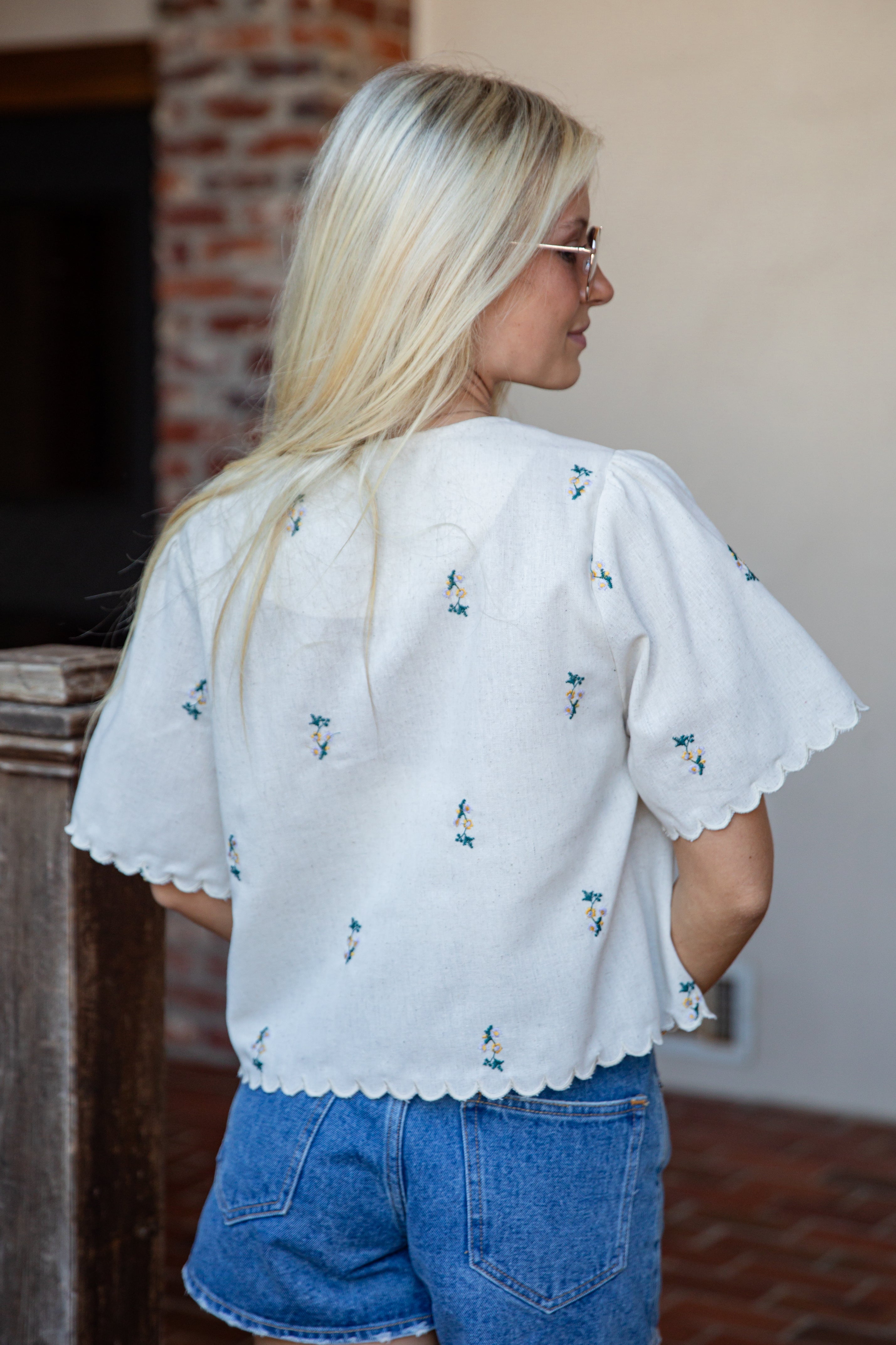 A woman with long blonde hair is shown from the back wearing a white short-sleeve blouse with small blue floral embroidery and scalloped edges, paired with blue denim shorts.