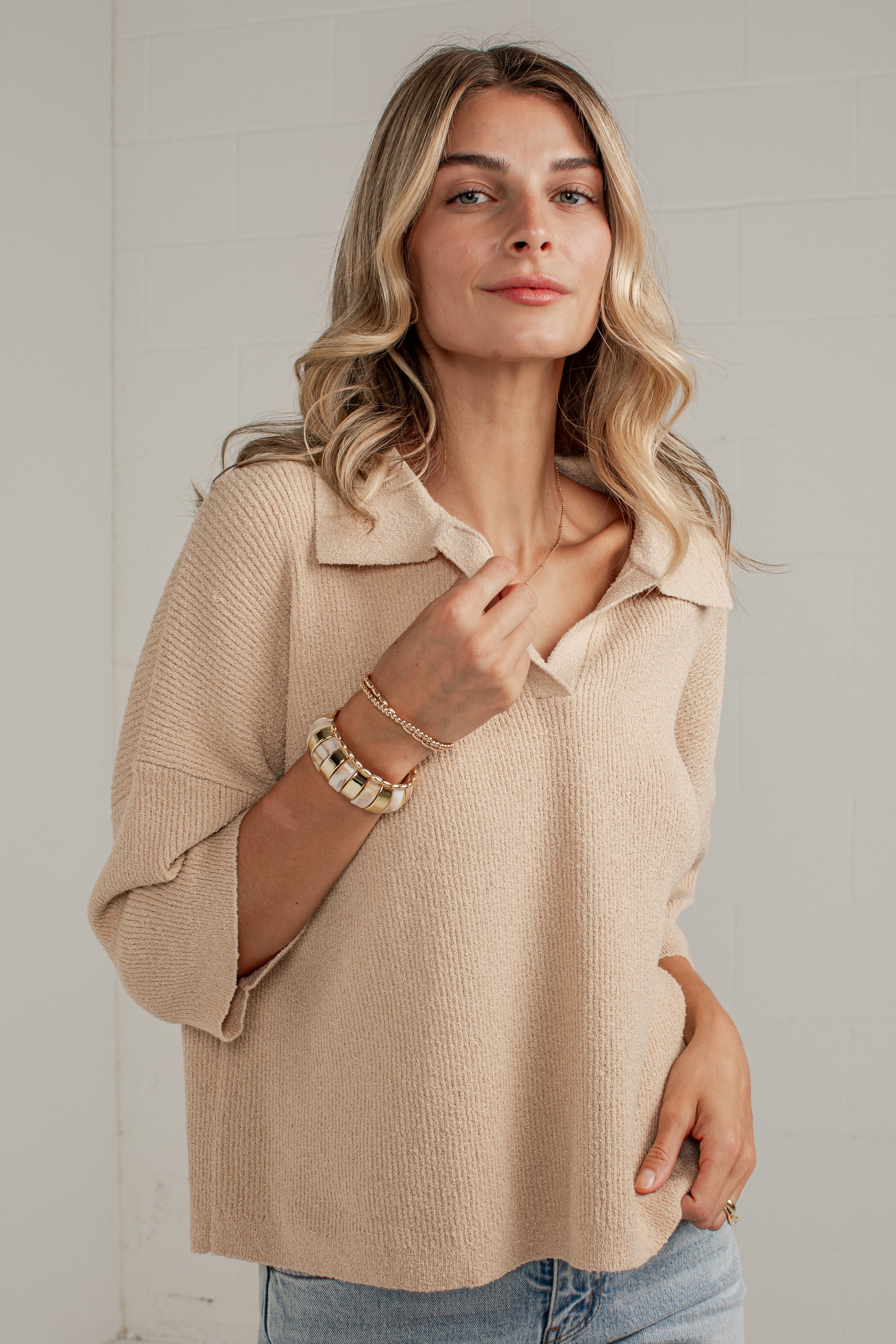A woman wearing a beige knit polo shirt with three-quarter sleeves and a collar, paired with light blue jeans, standing against a light gray brick wall.