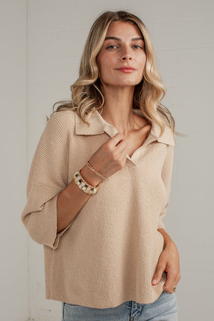 A woman wearing a beige knit polo shirt with three-quarter sleeves and a collar, paired with light blue jeans, standing against a light gray brick wall.