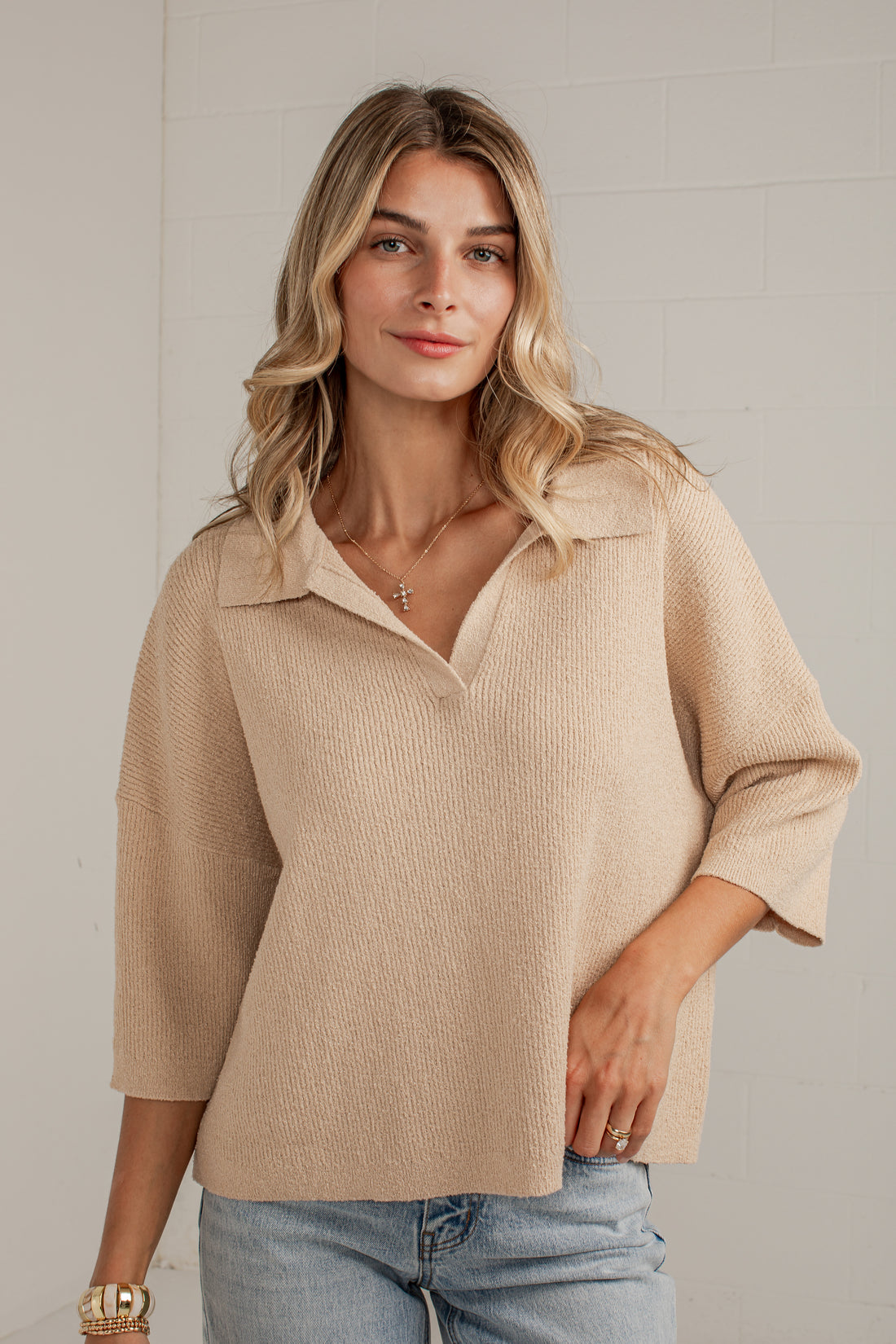 A woman wearing a beige textured knit polo shirt with three-quarter sleeves and a split collar, paired with light blue jeans, standing against a light gray brick wall.