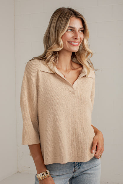A woman wearing a beige ribbed knit polo shirt with three-quarter sleeves and a split collar, paired with light-wash blue jeans, standing against a white brick wall.