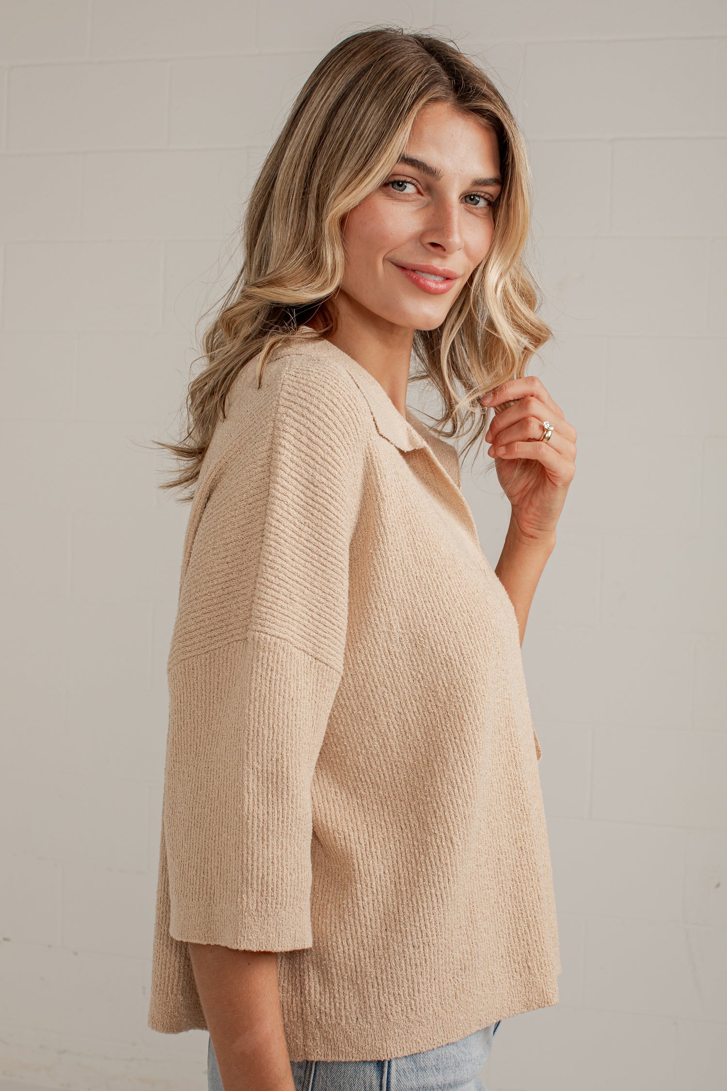 A woman wearing a textured, light beige knit top with three-quarter sleeves, shown from a side-back angle against a light gray brick wall.