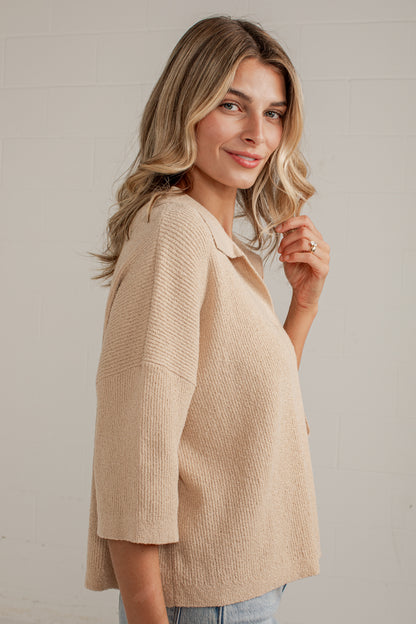 A woman wearing a textured, light beige knit top with three-quarter sleeves, shown from a side-back angle against a light gray brick wall.