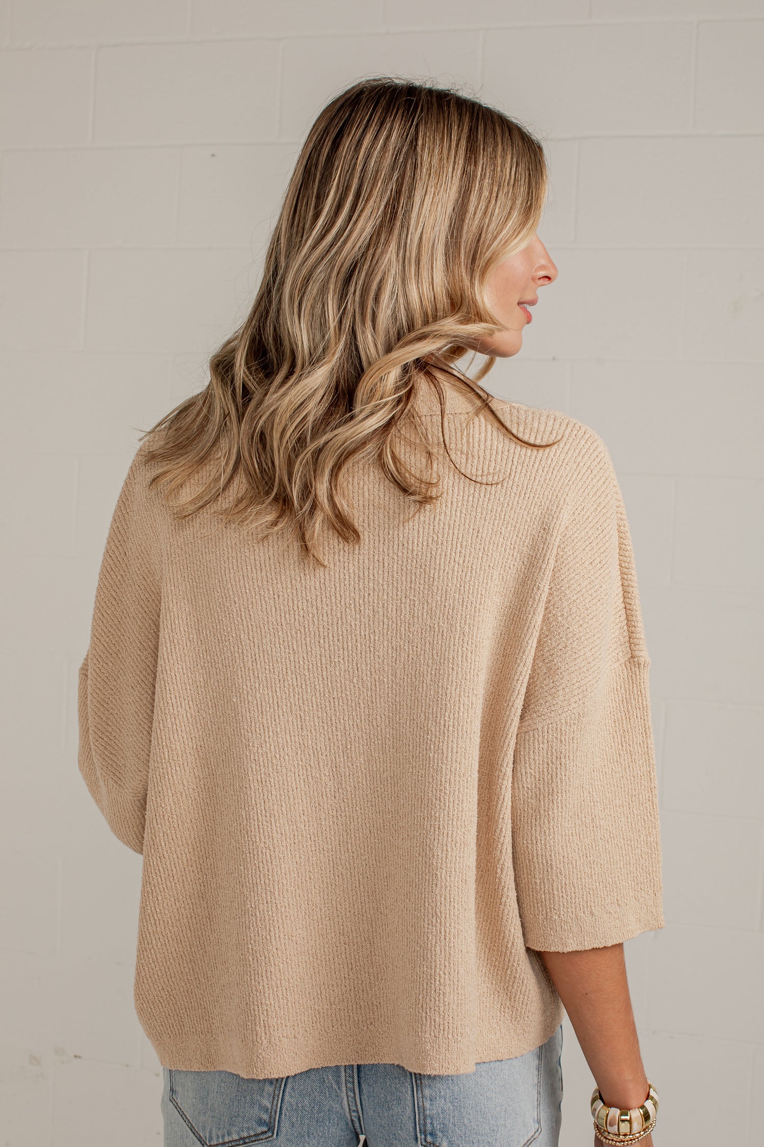 A woman with blonde wavy hair is shown from the back wearing a beige knit sweater and light blue jeans against a white brick wall.