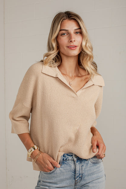 A woman wearing a beige ribbed knit polo sweater and light blue jeans, standing against a white brick wall.