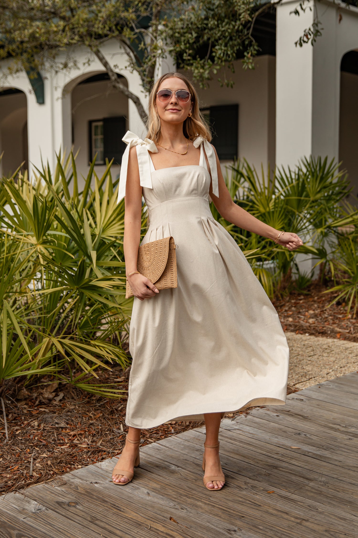 Woman in a white dress standing on a wooden deck with greenery and a building in the background