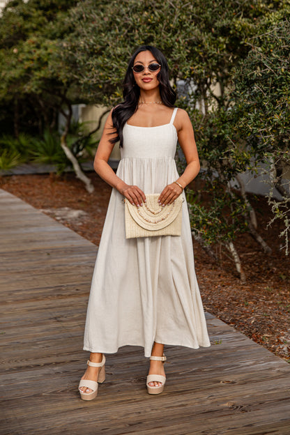 Woman in a white dress holding a beige handbag on a wooden path with greenery.