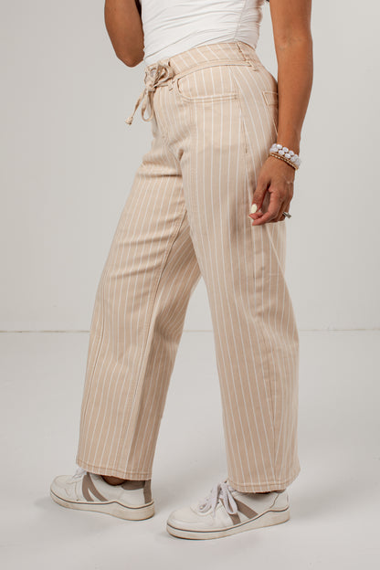 A person wearing beige wide-leg pinstripe pants with a drawstring waist, styled with a white top and white sneakers, shown from the waist down against a light gray studio background.