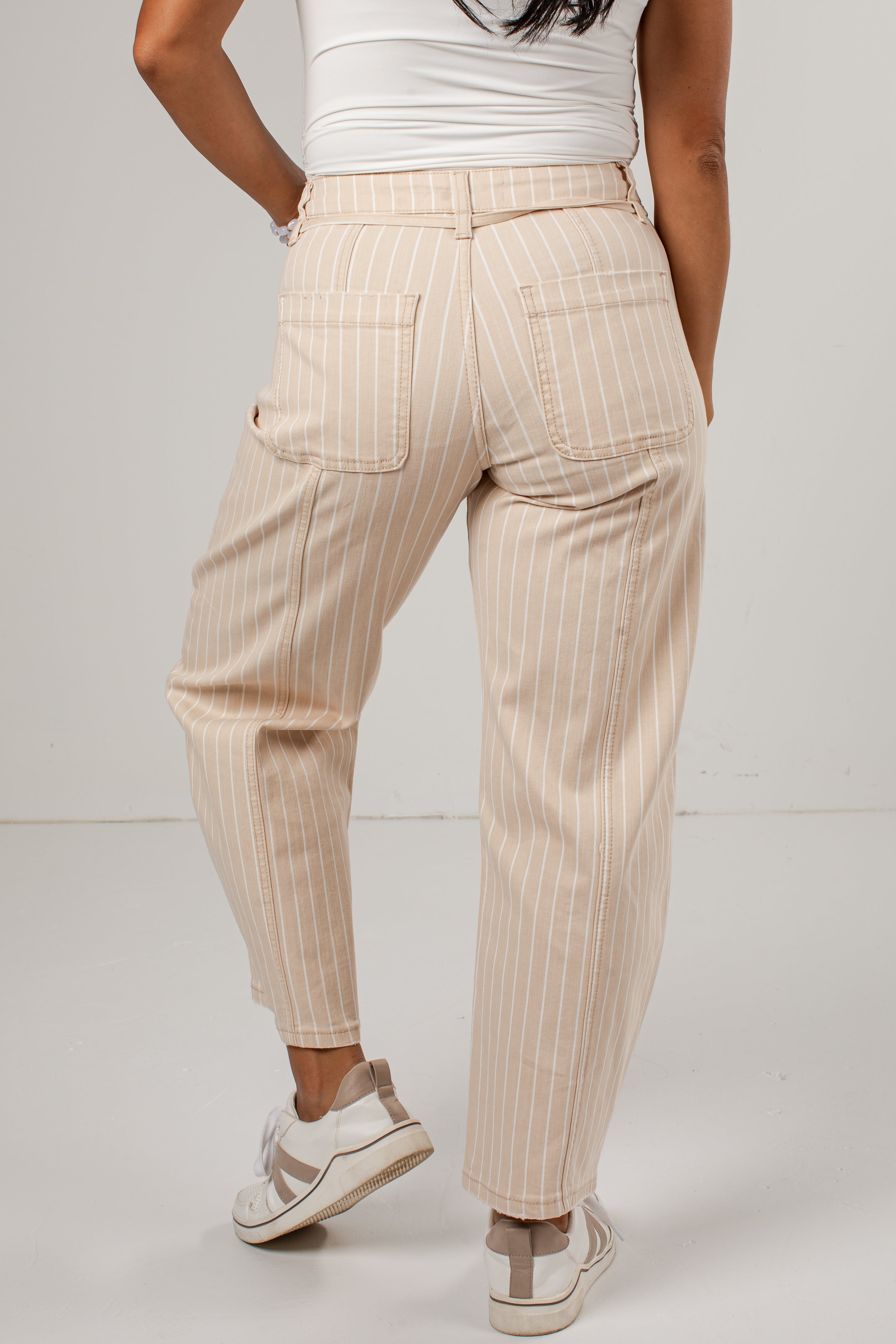 A person shown from the back wearing light beige pinstriped pants with a white top and white sneakers, photographed against a plain light gray studio background.