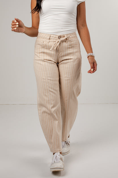 A model wearing beige pinstriped drawstring pants, a white ribbed tank top, and white sneakers, walking against a light gray studio background.