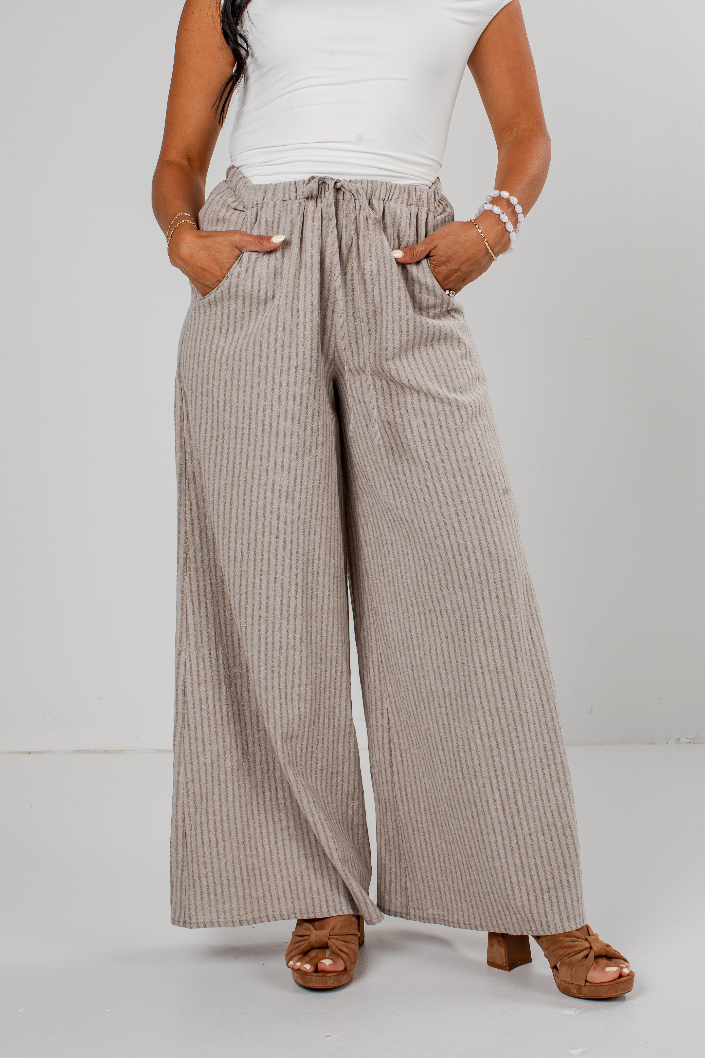 A model wearing high-waisted, wide-leg trousers with vertical taupe and white pinstripes, paired with a white top and brown heeled sandals.