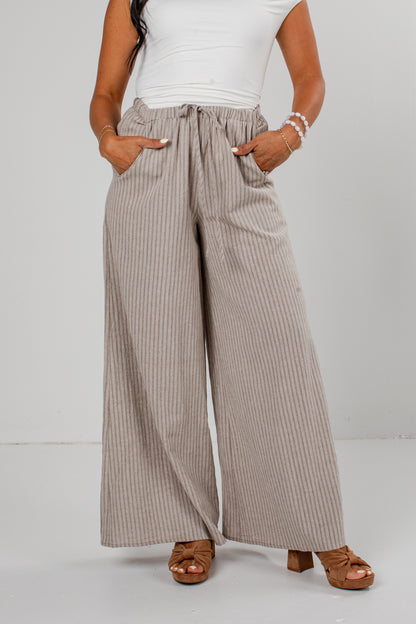 A model wearing high-waisted, wide-leg trousers with vertical taupe and white pinstripes, paired with a white top and brown heeled sandals.