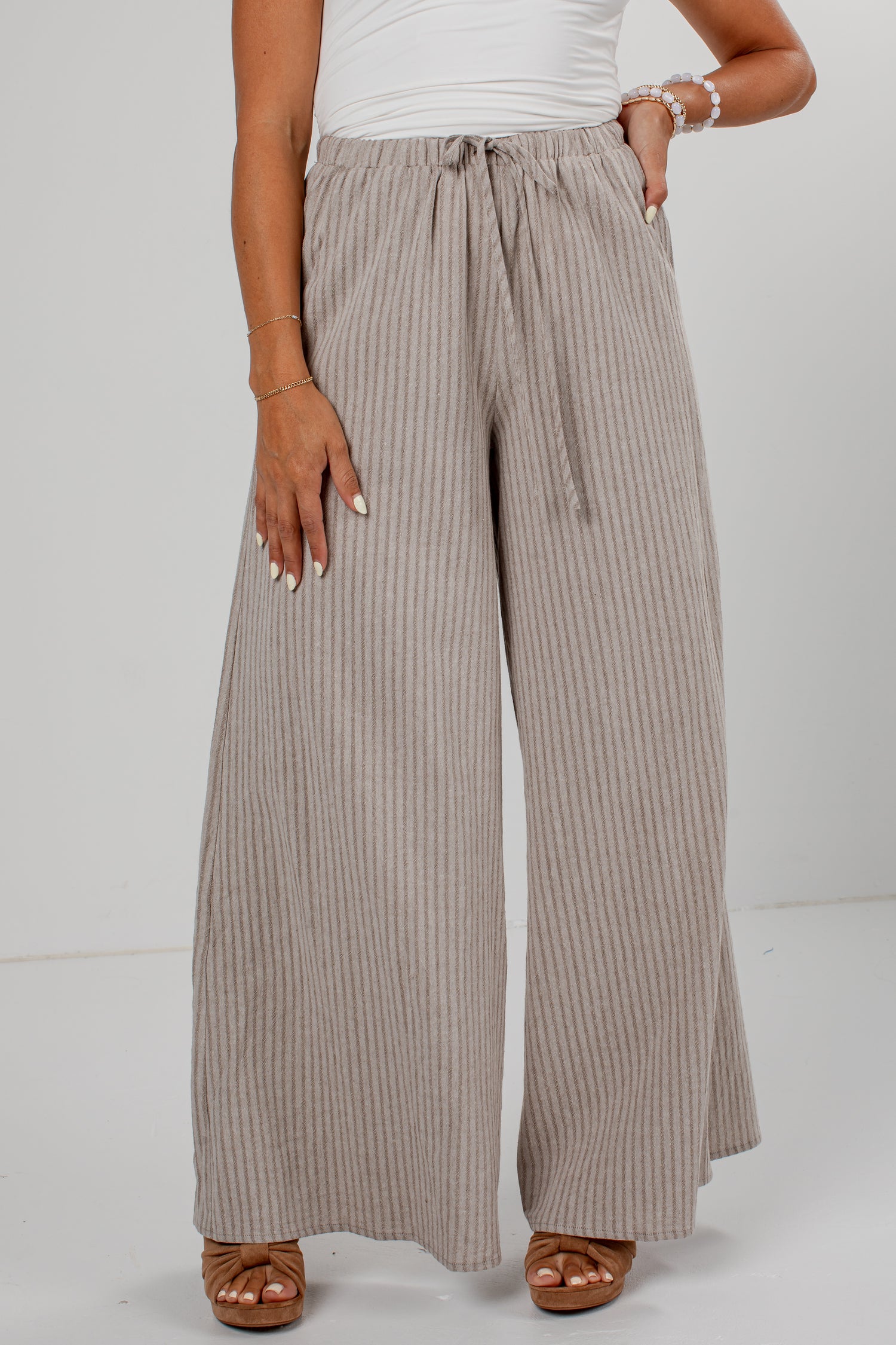 A person wearing wide-leg pleated pants in a taupe-beige color with thin vertical stripes, paired with a white top and brown sandals, shown from the waist down against a light gray studio background.