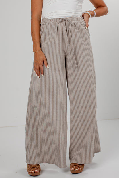 A person wearing wide-leg pleated pants in a taupe-beige color with thin vertical stripes, paired with a white top and brown sandals, shown from the waist down against a light gray studio background.