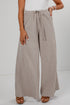 A person wearing wide-leg pleated pants in a taupe-beige color with thin vertical stripes, paired with a white top and brown sandals, shown from the waist down against a light gray studio background.