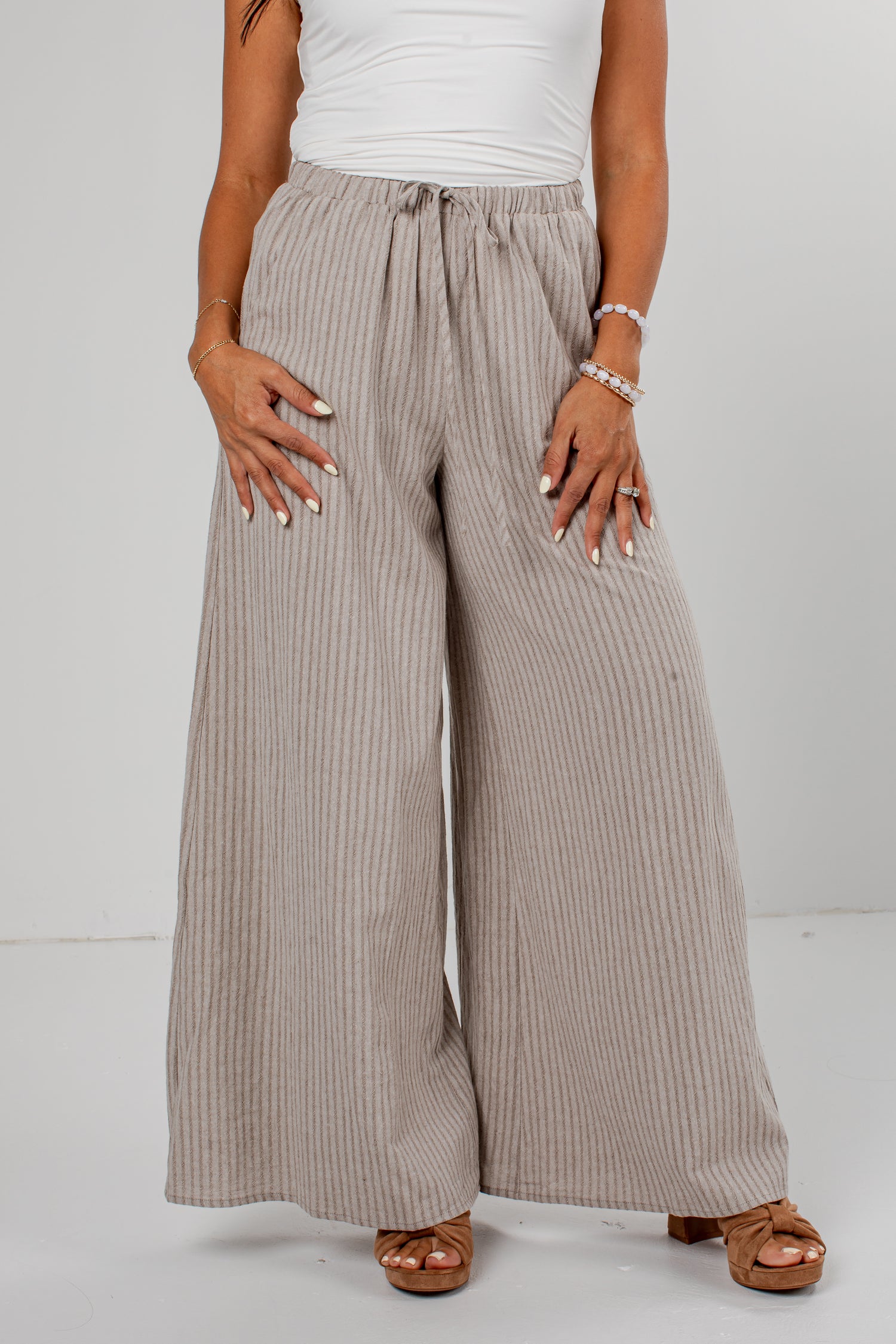 A person wearing wide-leg pleated pants in a light taupe-beige color, styled with a white top and brown sandals against a plain light gray studio background.