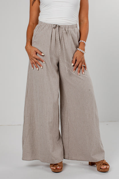 A person wearing wide-leg pleated pants in a light taupe-beige color, styled with a white top and brown sandals against a plain light gray studio background.